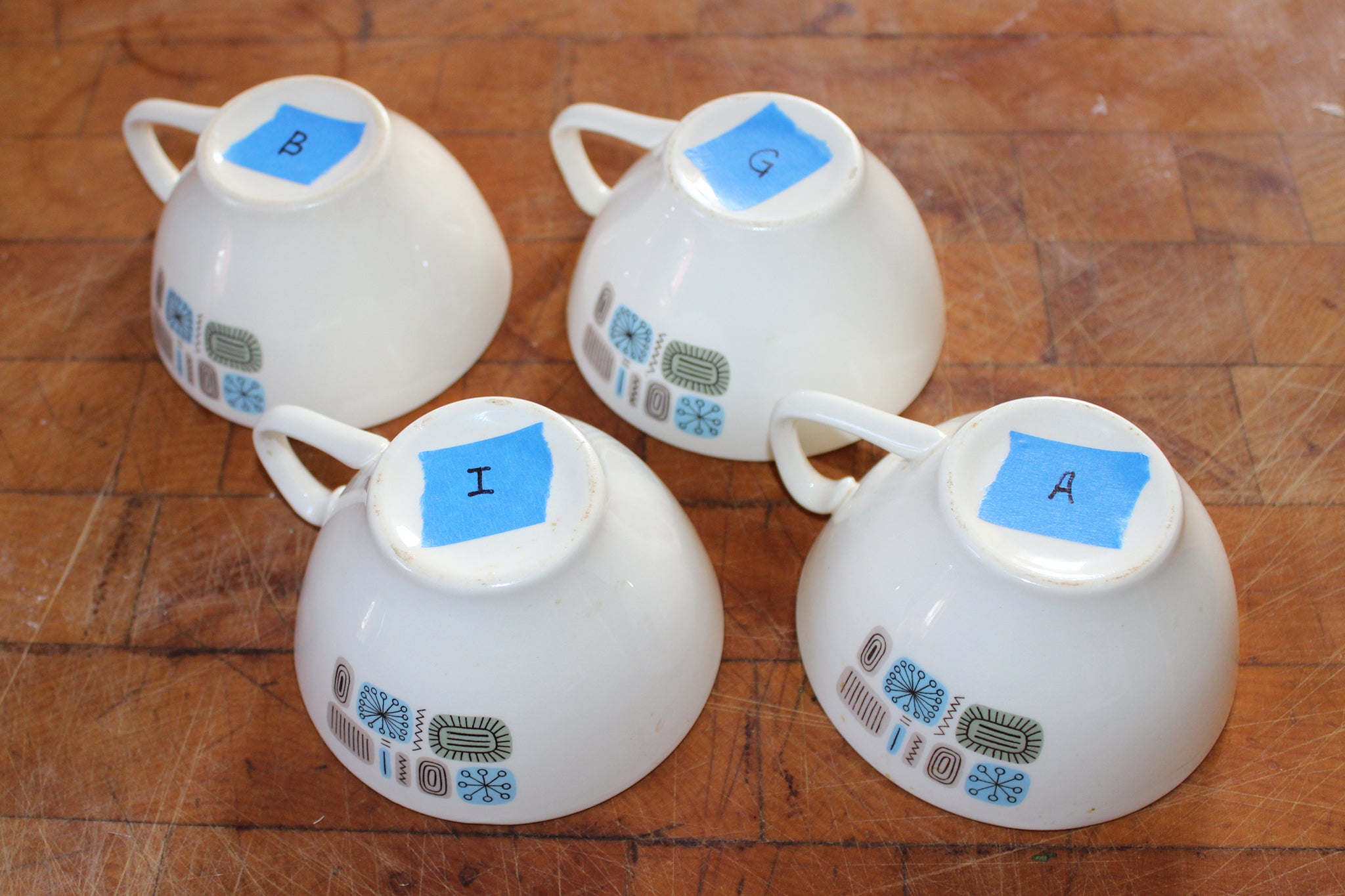 Four porcelain coffee cups sit upside down on a table. They are each labeled with a different letter on blue painter's tape.