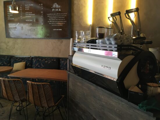 The espresso coffee machine at Cafe Piha, a La Marzocco with a white and chrome exterior, long metal handles for pulling shots, and cups stacked on top.