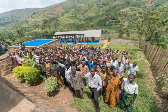 A large group picture of the Ruli Mountain coffee farmers of Rwanda. There are dozens of men and women, both young and old; some are in traditional Rwanda dress and others in suits and Western attire. Lots of wear hats. They are raising their right hands in greeting and smiling. Behind the group, which is outside on the grass, is a small mountain and a covered porch.