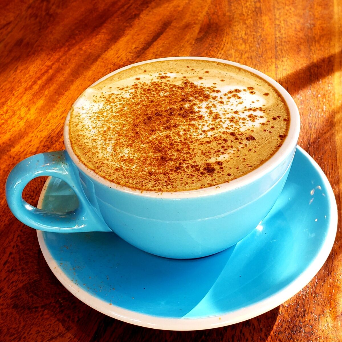 A sky blue ceramic mug and saucer is filled with a coffee beverage and sprinkled with cinnamon. It sits on a wood table top.