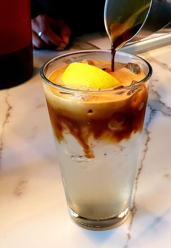 A tall glass of sparkling water is topped with a lemon wedge. Espresso coffee is being poured on top from a metal pitcher.
