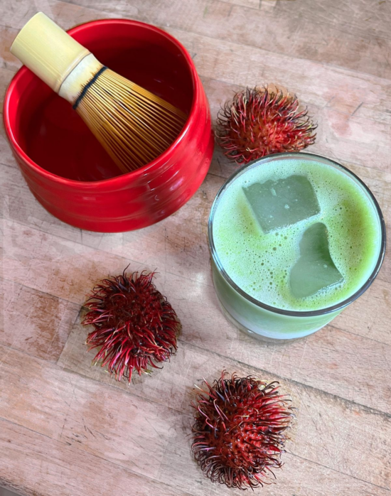An photo of a red matcha bowl with a bamboo matcha whisk inside it. There is a glass of iced matcha beside the bowl. Scattered on the table are three small rambutan fruits, still whole.