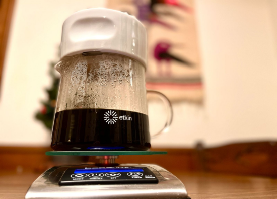 The Dripper on top of a scale with coffee making through it. The etkin symbol (similar to a sun, identical to the raised lines of the inside of the dripper) and the world etkin is printed on the side of the clear glass carafe.