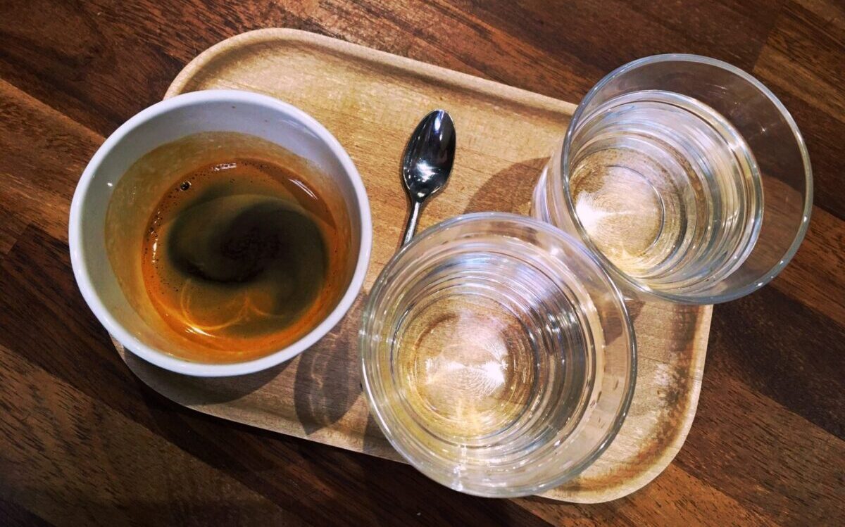 An espresso coffee in white cup. There are are two small water glasses and an espresso coffee spoon on the wooden tray.