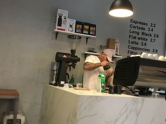 A barista in a ball cap pours a latte behind the bar at The Troubleshoot. The menu is pasted directly on the white wall behind. White shelves hold coffee bags and a chemex and other devices. The bar looks like white and gray marble.