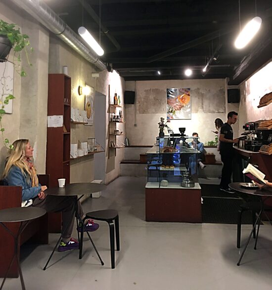 Customers sit inside Acid #1 while baristas make drinks. There is abstract art on the walls, small black tables with wooden benches on a concrete floor, and glass pastry case. The ceiling is black with bright industrial lighting.