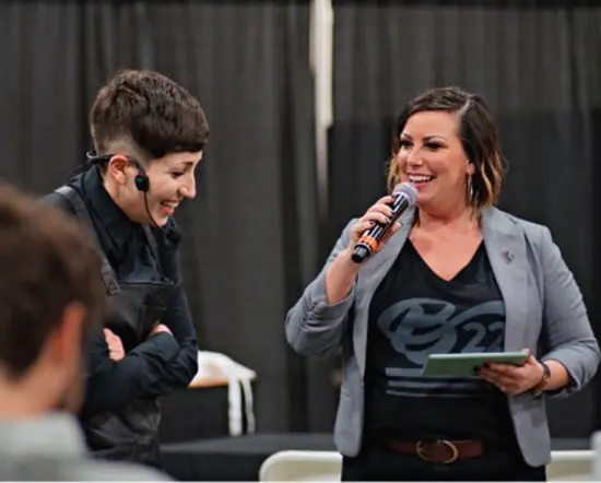 Anna holds a microphone and smiles while talking to a smiling competitor in a headset.